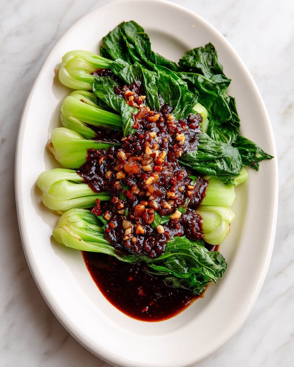 A white plate holds one layer of bright green steamed leafy greens arranged neatly, with visible long stems extending from one side. The greens have a fresh and soft texture, slightly wilted from cooking. On top, there is a thick dark brown sauce with a shiny surface that spreads over the middle portion of the leaves, containing small chunks of pale garlic or nuts adding rough texture and contrast. Some sauce gently flows off the side of the greens onto the white plate, highlighting the dish's rich and savory appearance. The background is a white marbled texture. photo taken with an iphone --ar 4:5 --v 7