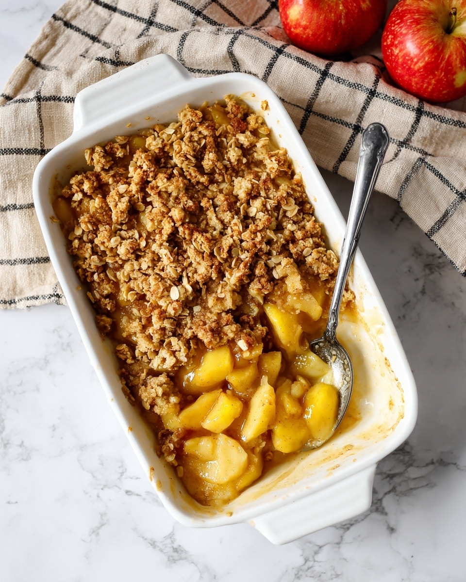 A white square baking dish filled with a warm apple crumble dessert that has two layers: the bottom layer consists of soft, chunky apple pieces in golden caramel sauce, and the top layer is a rough, crumbly oat topping with a golden-brown color, sprinkled unevenly, showing bits of apple peeking through. A spoon rests inside the dish on the right side, partially submerged in the apple filling. The dish is placed on a white marbled surface with a beige and black checkered cloth partly visible behind it and two red apples in the background on the right side. photo taken with an iphone --ar 4:5 --v 7