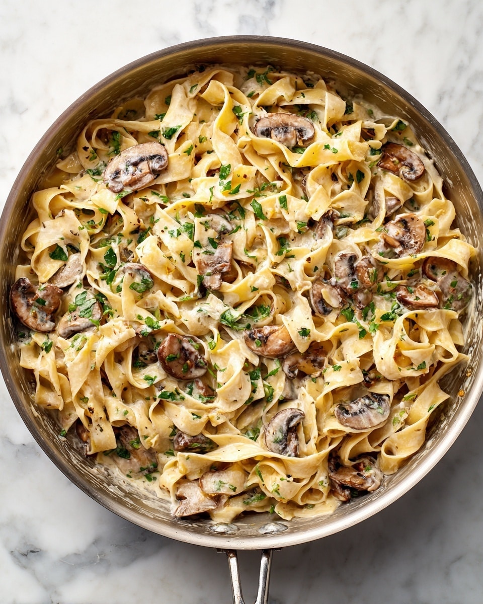 The image shows a creamy pasta dish with wide, flat noodles mixed with sliced mushrooms in a white sauce. The noodles are soft, pale beige, and slightly curled, evenly spread across the pan with mushrooms scattered throughout, adding dark brown and light gray tones. Small green herb pieces are mixed lightly into the sauce, giving a fresh contrast. The creamy sauce coats the noodles and mushrooms smoothly, with a few slight golden spots where the sauce has cooked. The pan is placed on a white marbled surface. Photo taken with an iphone --ar 4:5 --v 7