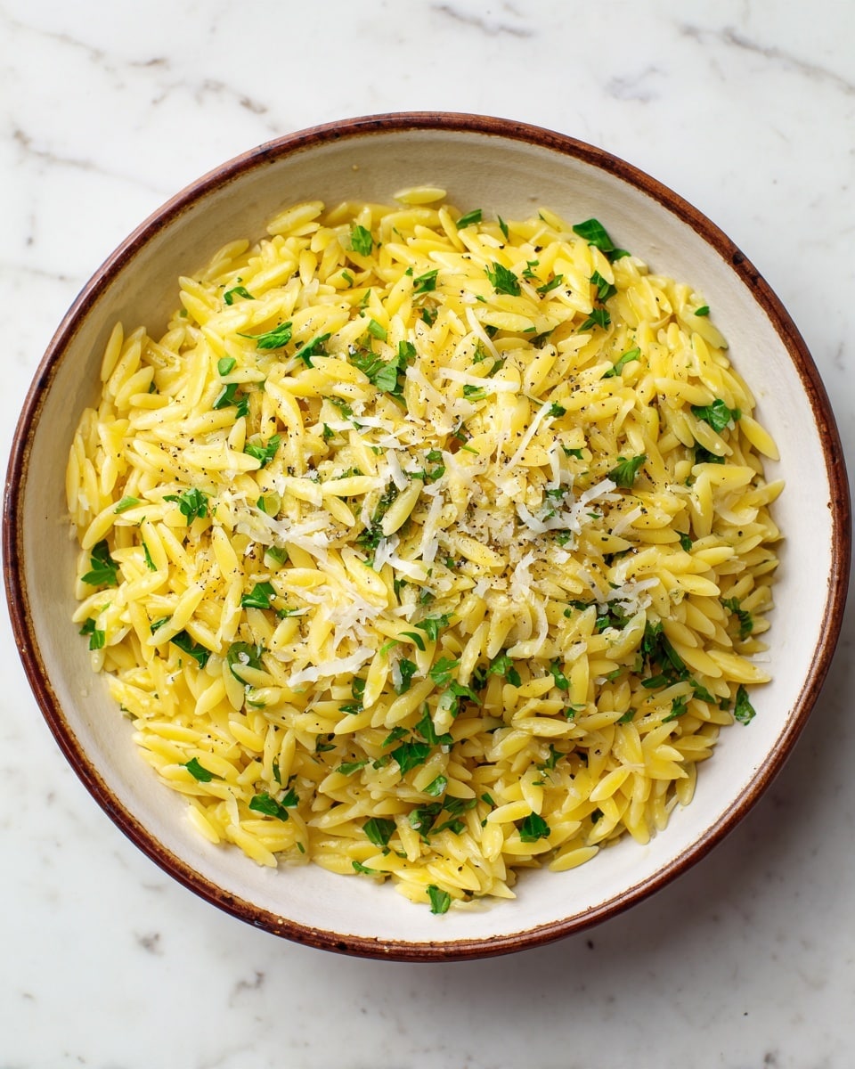 A close-up view of a bowl filled with yellow cooked orzo pasta, mixed evenly with small chopped green herbs, and topped with finely grated white cheese and a light sprinkle of black pepper. The orzo is glossy and looks soft, piled high in a white bowl with a brown rim. The background is a white marbled surface, creating a clean, fresh setting. photo taken with an iphone --ar 4:5 --v 7