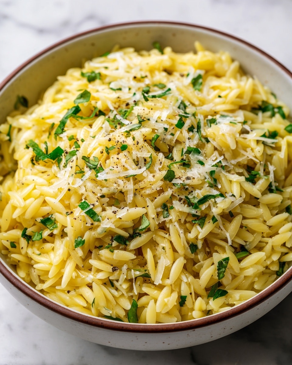 A close-up view of a round white plate with a rustic brown rim filled with creamy orzo pasta. The orzo is cooked and mixed with small green chopped herbs, likely parsley, and topped with a light sprinkle of grated cheese and cracked black pepper. The orzo has a glossy, smooth texture with a soft golden-yellow color. The dish sits on a white marbled surface. Photo taken with an iphone --ar 4:5 --v 7