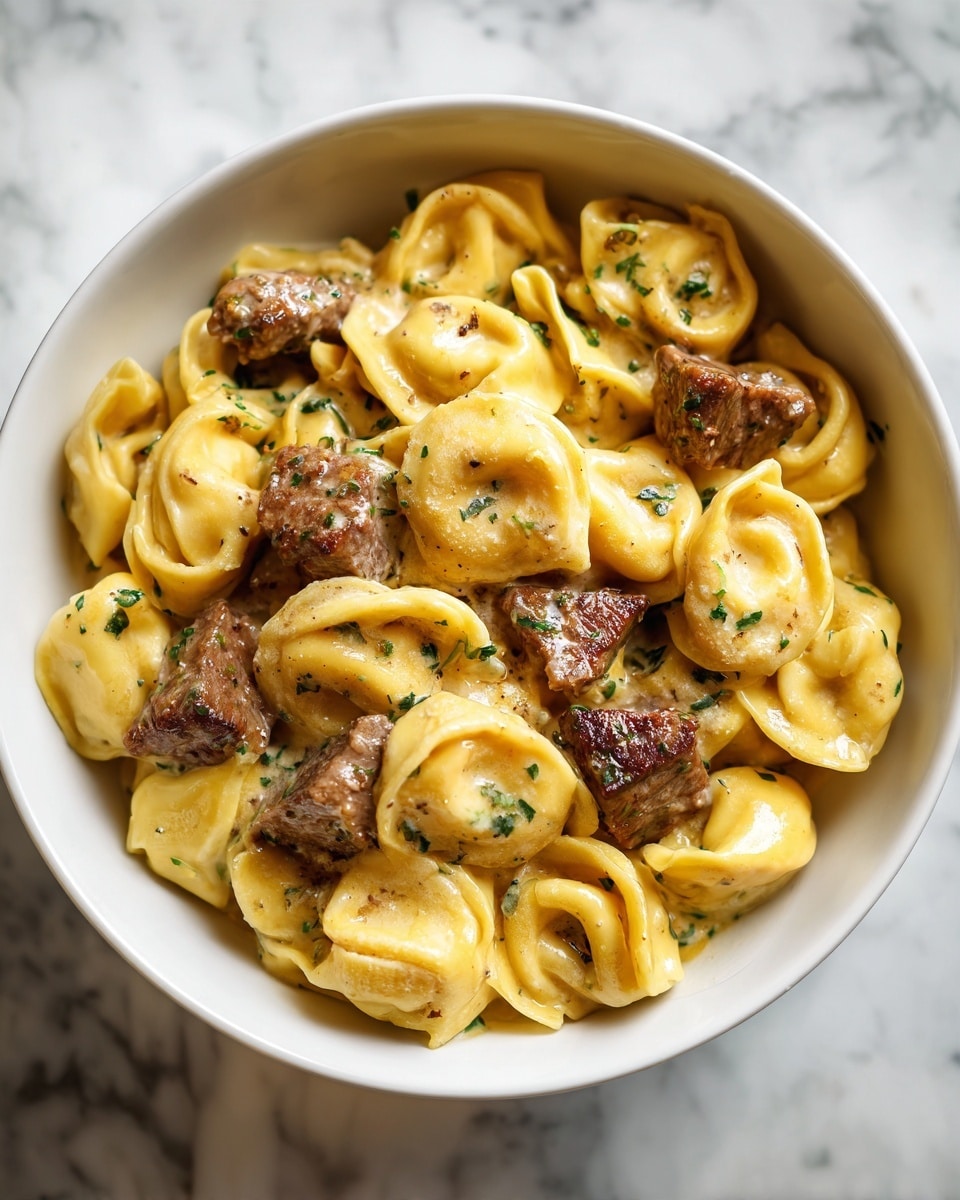 The image shows a white bowl filled with two main layers: the bottom layer is creamy yellow tortellini pasta with a smooth texture and a slightly glossy finish; on top are several chunks of browned beef, textured and cooked with visible searing marks. The dish is sprinkled evenly with small green herb pieces and cracked black pepper, adding color contrast. The background is a white marbled surface. Photo taken with an iphone --ar 4:5 --v 7